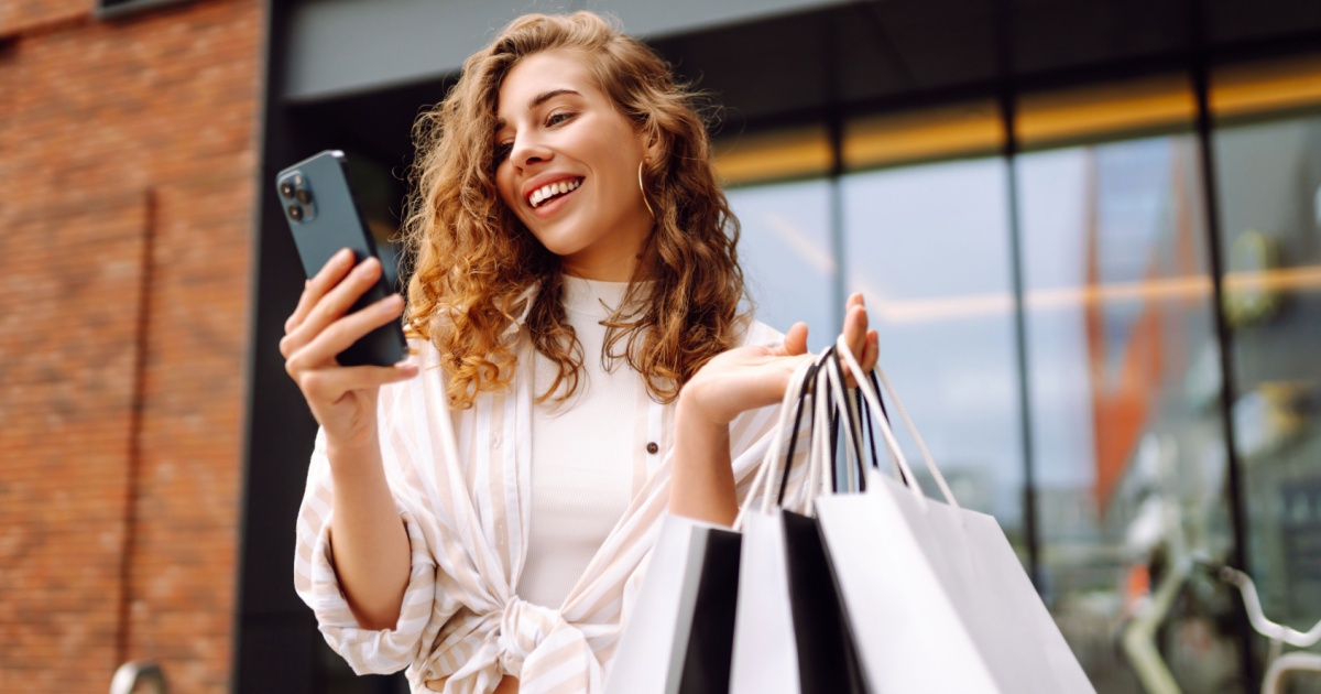 A imagem mostra uma mulher sorrindo enquanto segura várias sacolas de compras em uma das mãos e um smartphone na outra. Ela está ao ar livre, em frente a um estabelecimento comercial moderno, com janelas grandes ao fundo. A mulher tem cabelos cacheados e ruivos, e veste uma blusa branca com listras. Ela parece feliz e satisfeita com suas compras, sugerindo uma experiência de compra positiva, possivelmente em um ambiente de varejo ou retailtech (compras, varejo, celular, sacolas, mulher, ruiva, smartphone, retailtechs, consumidor)