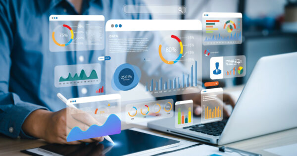 Pessoa interagindo com gráficos digitais flutuantes em um ambiente de trabalho, representando análise de dados e inovação tecnológica, com laptop e tablet ao fundo, mercado de TI