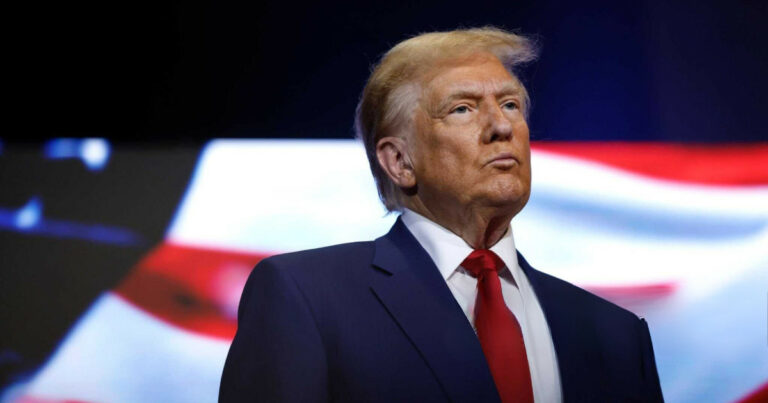 Donald Trump, presidente dos Estados Unidos, em destaque. Ele está usando um terno azul escuro, camisa branca e gravata vermelha, com uma expressão séria e olhar fixo. Ao fundo, há um padrão desfocado em EUA vermelho, branco e azul, remetendo às cores da bandeira americana. A imagem transmite formalidade e autoridade (ordem executiva, ações, apple, tarifas)