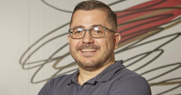Foto de Everson Ramos, da Vórtx, sorrindo e posando com os braços cruzados. Ele usa óculos e uma camisa polo azul. O fundo apresenta um design abstrato com linhas em tons de vermelho e preto (projeto)