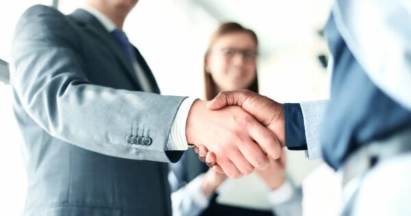 Dois executivos apertando as mãos em sinal de acordo, representando fusão e aquisição, com uma mulher ao fundo sorrindo (M&A, fusao, aquisicao, negocio, acordo, desafios, telecom, Populos, grupo, semestre, brasil, CEO, Catarin, engineering)