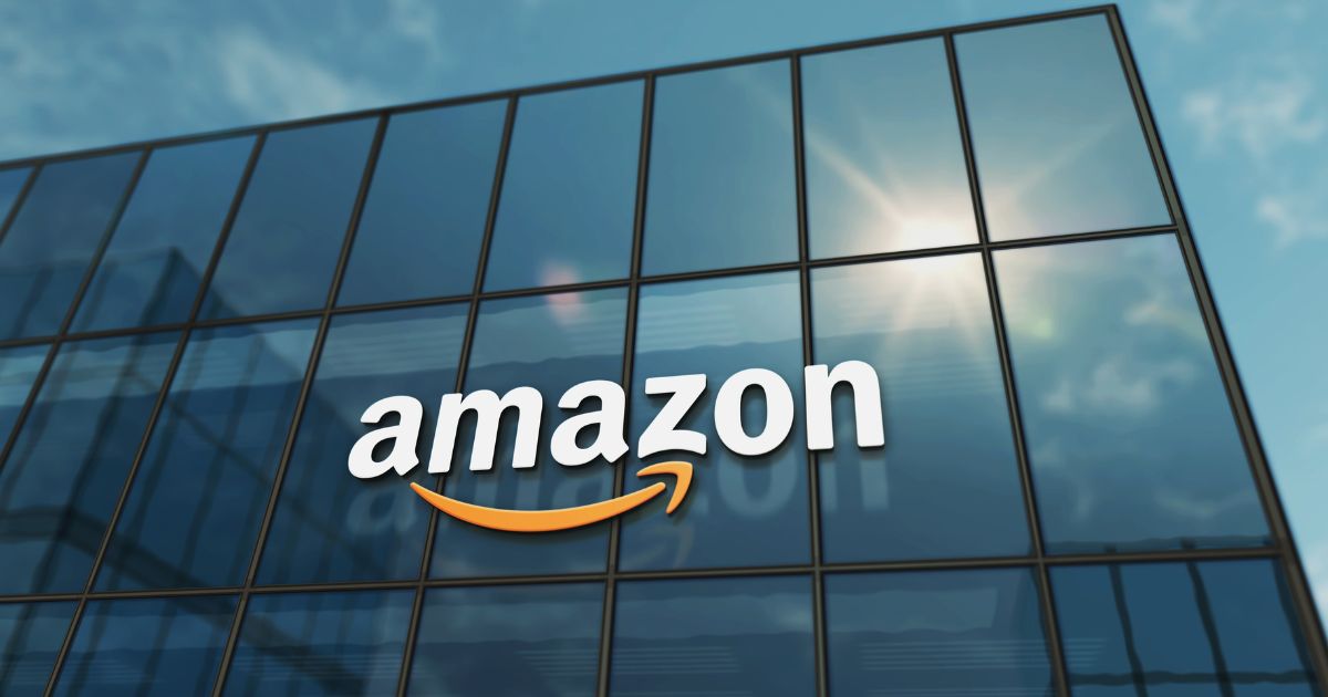 Fachada de um edifício moderno com grandes janelas de vidro refletindo o céu azul e o sol. O logótipo da Amazon, com letras brancas e a icónica seta amarela em forma de sorriso, está em destaque no vidro, contrastando com o fundo espelhado