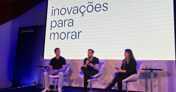 Três pessoas estão sentadas em cadeiras no palco de um evento com fundo iluminado, onde se lê 'inovações para morar'. À esquerda, um homem de camisa preta e calças jeans segura um microfone. No centro, outro homem, também com camisa preta e calças escuras, está falando ao microfone. À direita, uma mulher com roupa preta segura um microfone enquanto ouve. O logótipo da empresa 'QuintoAndar' é visível no canto superior esquerdo do palco, e há mesas pequenas com garrafas de água ao lado dos participantes