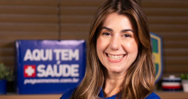 Priscila Braga, nova diretora de Digital e Inteligência de Vendas da Pague Menos, sorrindo em um ambiente corporativo. Ao fundo, um cartaz azul com os dizeres 'Aqui tem saúde' reforça a identidade da empresa