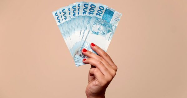 Imagem ilustrativa destacando várias cédulas de 100 reais sendo seguradas por uma mão feminina, representando conceitos como eConsignado, crédito consignado ou finanças pessoais, em um fundo bege uniforme (PaGol) portabilidade