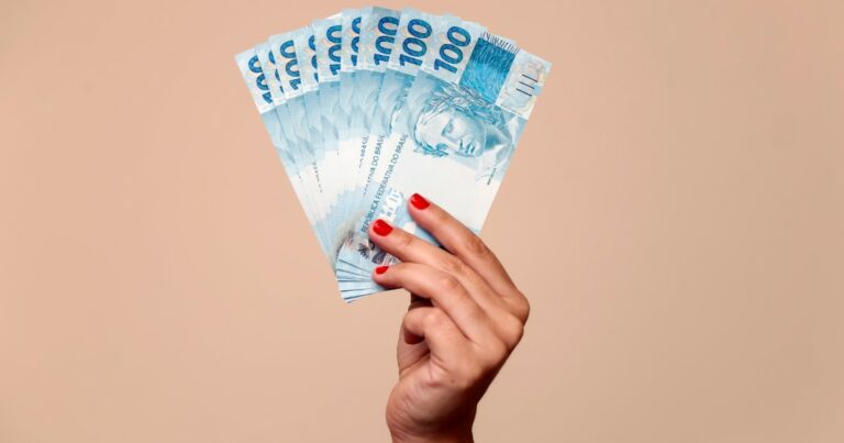 Imagem ilustrativa destacando várias cédulas de 100 reais sendo seguradas por uma mão feminina, representando conceitos como eConsignado, crédito consignado ou finanças pessoais, em um fundo bege uniforme (PaGol) portabilidade