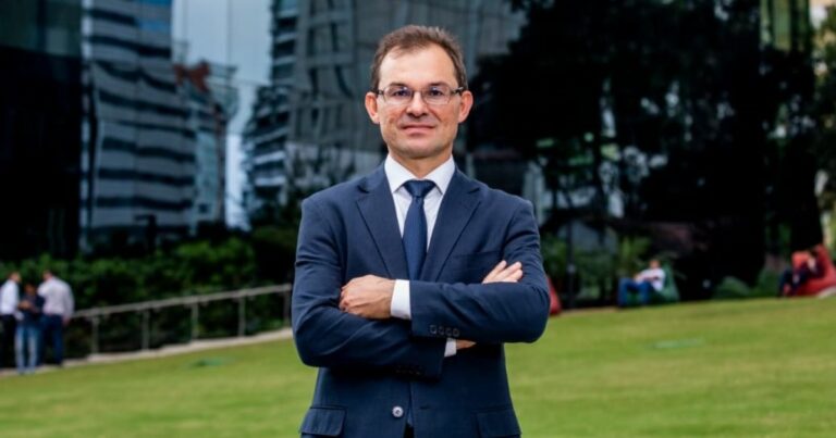 Imagem de Marcos Ferrari, presidente-executivo da Conexis, em um ambiente externo. Ele está usando um terno azul escuro com gravata, de braços cruzados, transmitindo uma postura confiante e profissional. Ao fundo, há um gramado verde, árvores e edifícios modernos, criando um cenário corporativo ao ar livre (Open Gateway, telecom)
