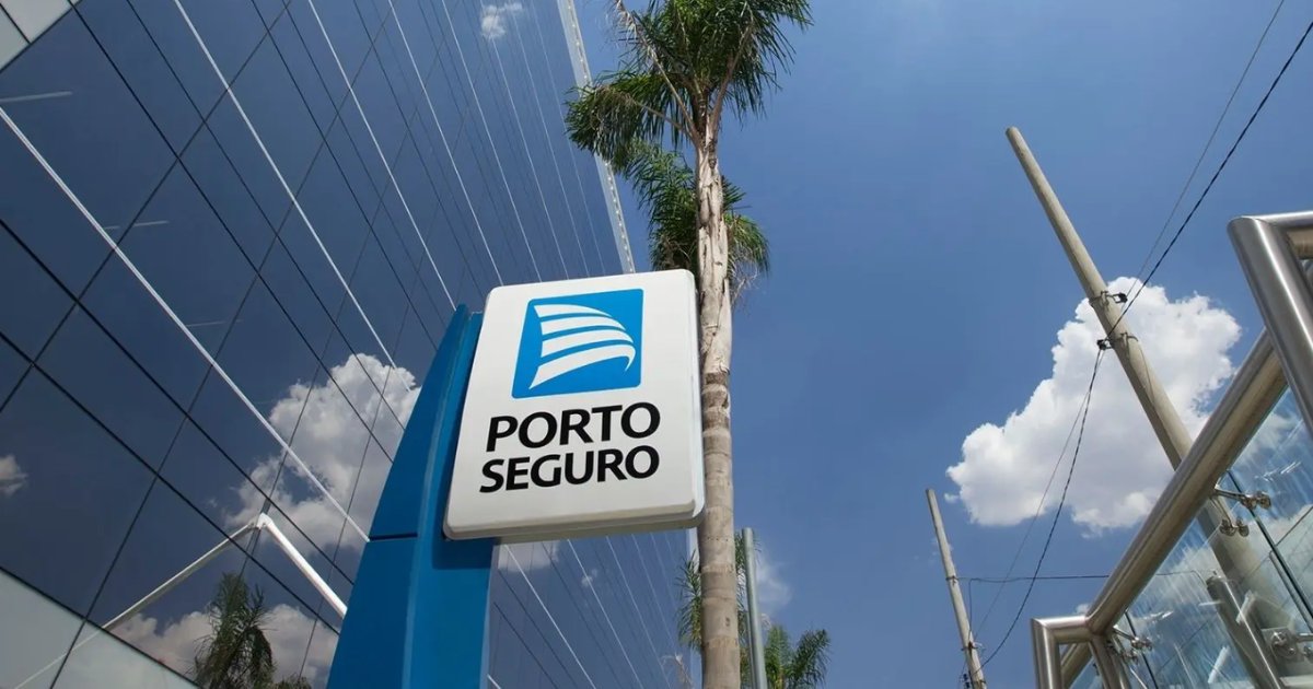 Placa com o logotipo da Porto Seguro em frente a um prédio espelhado, com palmeiras e céu azul com nuvens ao fundo.