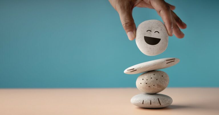 Mão posicionando uma pedra com rosto sorridente no topo de uma pilha de pedras decoradas com desenhos simples. O fundo azul claro e a superfície bege criam uma composição minimalista, simbolizando equilíbrio, bem-estar e felicidade (saúde mental)