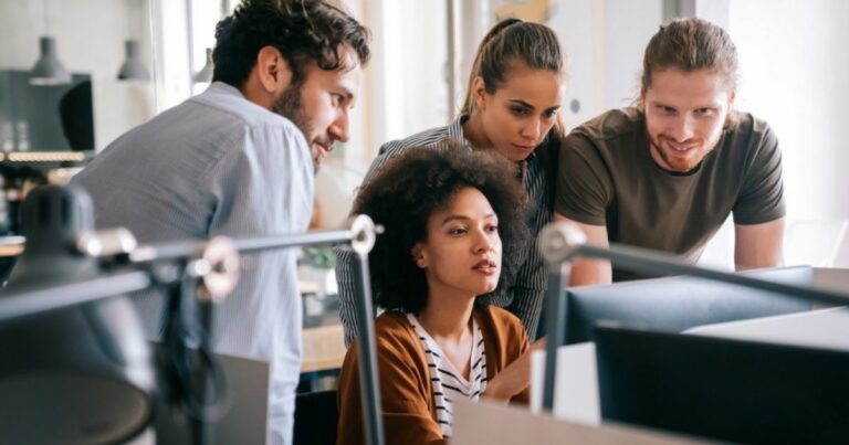A imagem mostra um grupo de quatro jovens profissionais reunidos ao redor de um computador em um ambiente de escritório moderno. A mulher no centro, de cabelo cacheado, parece estar explicando algo na tela enquanto os outros três, um homem de barba, uma mulher de cabelo preso e outro homem de cabelo longo, observam atentamente. O ambiente tem um design contemporâneo, com luminárias pendentes e um espaço bem iluminado. A cena transmite colaboração e trabalho em equipe (positivo tecnologia, positivo S+, especiali)