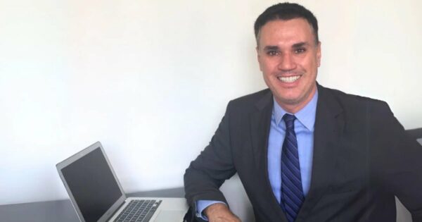 A imagem mostra Franklin Tomich, fundador da Accordia, sentado à mesa com um laptop ao lado. Ele veste um terno escuro, camisa azul e gravata listrada, transmitindo um visual profissional. Com um sorriso confiante, ele olha diretamente para a câmera. O fundo é simples, com uma parede branca, destacando o ambiente corporativo e minimalista da foto (Franklin Tomich, Accordia, risco, planejamento)