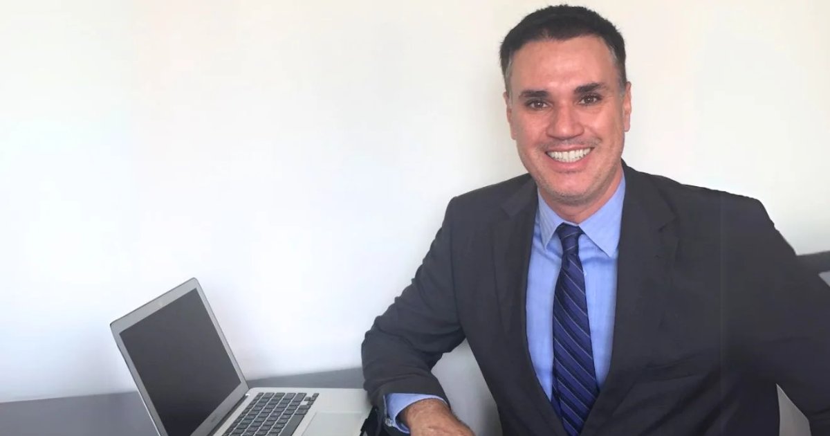 A imagem mostra Franklin Tomich, fundador da Accordia, sentado à mesa com um laptop ao lado. Ele veste um terno escuro, camisa azul e gravata listrada, transmitindo um visual profissional. Com um sorriso confiante, ele olha diretamente para a câmera. O fundo é simples, com uma parede branca, destacando o ambiente corporativo e minimalista da foto (Franklin Tomich, Accordia, risco, planejamento)