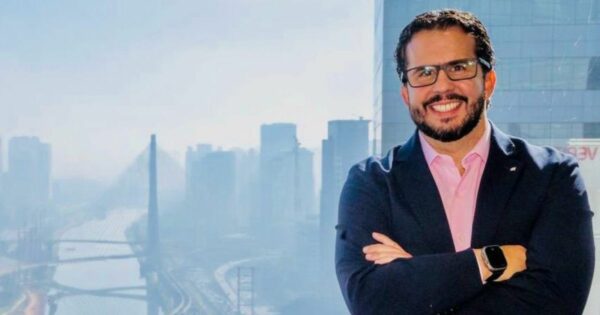 Homem sorridente com óculos e blazer azul posando em frente a uma janela com vista para a cidade de São Paulo, destacando a Ponte Estaiada e prédios ao fundo. Gustavo Leite, VP América Latina e Caribe da Cohesity. Foto: Divulgação
