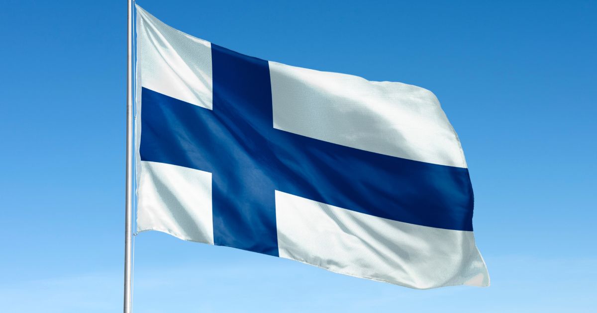 Uma bandeira da Finlândia tremula ao vento contra um céu azul claro. A bandeira tem um fundo branco com uma cruz azul escura no centro, estendendo-se horizontal e verticalmente. O tecido da bandeira parece ligeiramente ondulado devido ao vento.