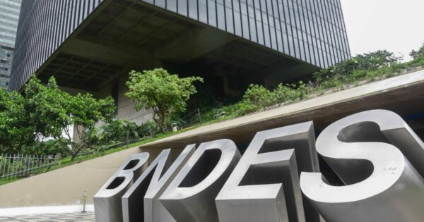 Fotografia da fachada do edifício do BNDES (Banco Nacional de Desenvolvimento Econômico e Social), com um letreiro metálico grande exibindo a sigla "BNDES" em destaque no primeiro plano. O prédio moderno, com arquitetura robusta e linhas retas, está cercado por áreas verdes e árvores. A perspectiva da foto enfatiza a grandiosidade da construção, com um céu nublado ao fundo 202