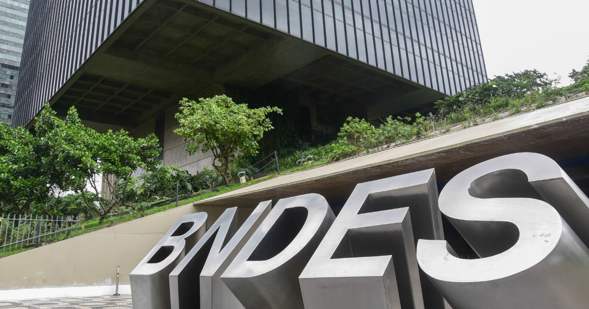 Fotografia da fachada do edifício do BNDES (Banco Nacional de Desenvolvimento Econômico e Social), com um letreiro metálico grande exibindo a sigla "BNDES" em destaque no primeiro plano. O prédio moderno, com arquitetura robusta e linhas retas, está cercado por áreas verdes e árvores. A perspectiva da foto enfatiza a grandiosidade da construção, com um céu nublado ao fundo 202