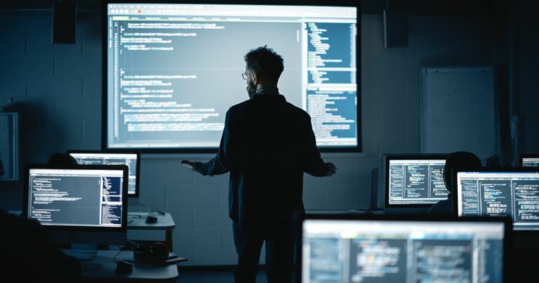 Uma sala escura de aula ou laboratório de pesquisa de informática com múltiplos monitores exibindo código de programação. No centro, um instrutor ou especialista em tecnologia, de costas para a câmera, gesticula enquanto explica algo para os alunos. Uma grande tela de projeção na parede exibe linhas de código, iluminando o ambiente com um brilho azulado. Os alunos, visíveis como silhuetas, estão concentrados em seus computadores. tec