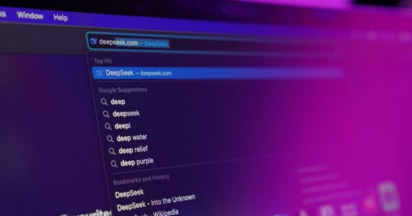 A imagem mostra um ecrã de computador com um navegador da web aberto, exibindo a barra de pesquisa onde foi digitado "deepseek.com". O site "DeepSeek" aparece como o principal resultado destacado. Abaixo, há sugestões de pesquisa do Google, incluindo termos como "deep", "deepseek", "deep water", "deep relief" e "deep purple". O ambiente apresenta um tom roxo e azul, refletindo uma iluminação neon, que cria um efeito futurista e tecnológico. O foco da imagem está na barra de pesquisa, enquanto o restante da interface do navegador está ligeiramente desfocado.