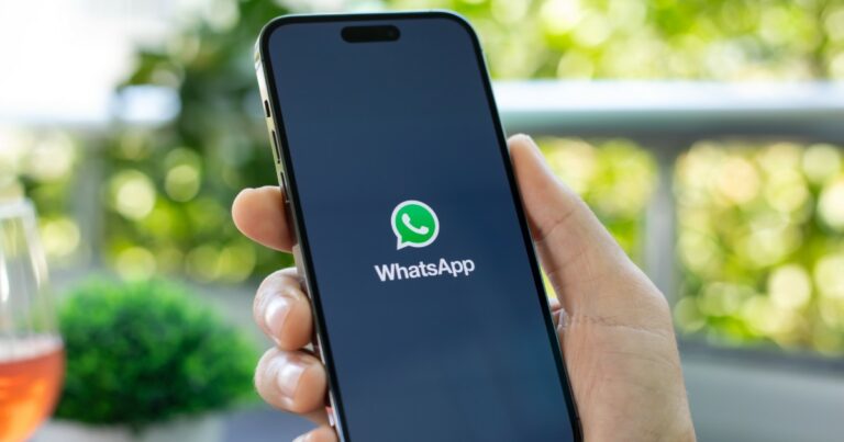 A imagem mostra uma mão segurando um smartphone com a tela ligada, exibindo a tela de carregamento do aplicativo WhatsApp. O logotipo verde e branco do WhatsApp está centralizado na tela preta, com o nome do aplicativo logo abaixo. O fundo da imagem está levemente desfocado, mostrando um ambiente ao ar livre com plantas verdes e um copo com bebida alaranjada ao lado, Exa (cade)