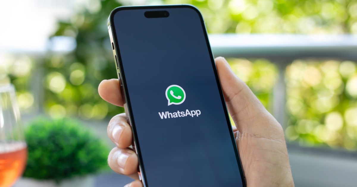 A imagem mostra uma mão segurando um smartphone com a tela ligada, exibindo a tela de carregamento do aplicativo WhatsApp. O logotipo verde e branco do WhatsApp está centralizado na tela preta, com o nome do aplicativo logo abaixo. O fundo da imagem está levemente desfocado, mostrando um ambiente ao ar livre com plantas verdes e um copo com bebida alaranjada ao lado, Exa (cade)