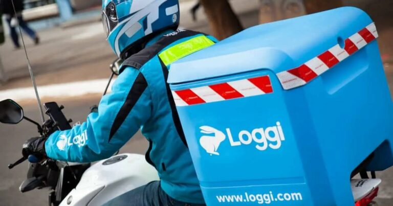 Motoboy da Loggi realizando uma entrega. Ele veste um uniforme azul com a logo da empresa e utiliza um capacete com detalhes brancos. A motocicleta possui um baú azul com o logotipo da Loggi e adesivos refletivos, reforçando a identidade visual da empresa de logística. A imagem transmite a agilidade e eficiência do serviço de entregas urbanas (loggi, motoboy, moto, entrega, logística, plataformas)