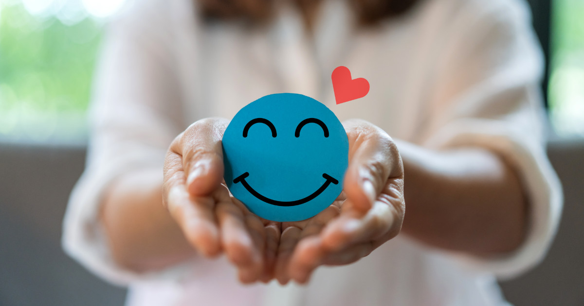A imagem mostra uma pessoa segurando delicadamente um círculo azul com um rosto sorridente desenhado, representando positividade e bem-estar. Acima, há um pequeno coração vermelho, reforçando a ideia de cuidado e saúde mental. O fundo é desfocado, dando destaque às mãos e ao símbolo de felicidade (NR-1)