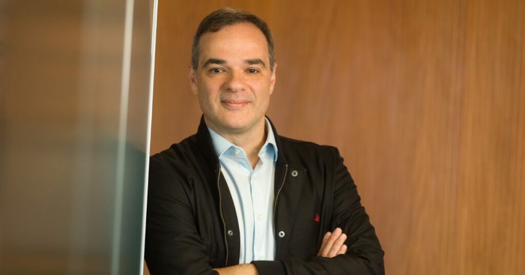 Felipe Ávila, da Livelo, sorri levemente com os braços cruzados, usando uma camisa azul clara e jaqueta preta, em frente a uma parede de madeira.