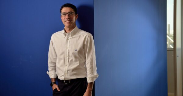 Imagem de Marcelo Mearim, CEO da Sofya, em pé diante de uma parede azul. Ele está sorrindo, veste uma camisa social branca de manga comprida com o logotipo discreto da marca na altura do peito e calça escura. Usa óculos e um relógio no pulso esquerdo. Ao fundo, há uma janela com vista externa parcialmente visível (saúde)