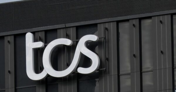 Um letreiro iluminado com as letras "tcs" em branco está fixado na fachada de um prédio moderno de cor escura. A estrutura do edifício apresenta janelas verticais e detalhes arquitetônicos minimalistas. O céu ao fundo está parcialmente nublado, refletindo sutilmente na superfície do letreiro (mulher)