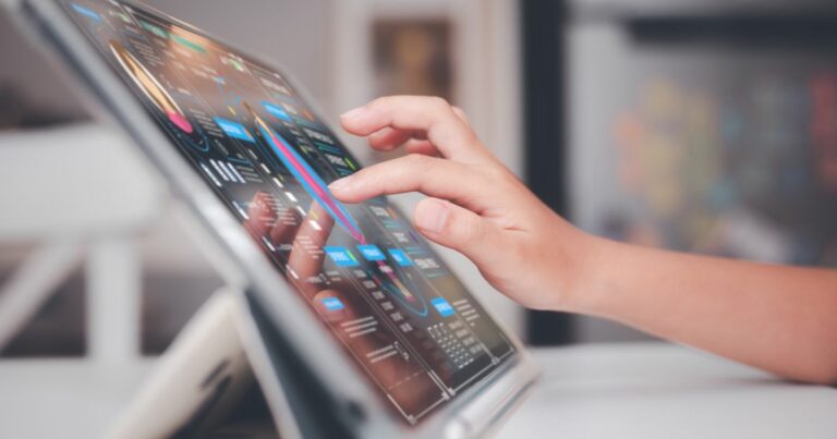 Mão interagindo com um tablet em um suporte, exibindo uma interface digital com gráficos, estatísticas e dados coloridos. A tela apresenta elementos visuais sofisticados, sugerindo uma análise de dados ou painel de controle digital. O fundo está desfocado, destacando o dispositivo e a interação do usuário (desafios)