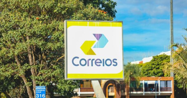 A imagem mostra uma placa dos Correios do Brasil em um ambiente urbano. O letreiro exibe o logotipo oficial da empresa, composto por duas setas estilizadas nas cores amarela e azul, apontando para a direita, junto com a palavra "Correios" escrita em letras azuis. A placa tem uma moldura metálica e fundo branco com detalhes em amarelo. No canto inferior esquerdo da imagem, há uma pequena placa azul com a inscrição "NORTE Comércio Local 316". Ao fundo, é possível ver árvores, um céu azul com algumas nuvens e edifícios com arquitetura típica de áreas comerciais e residenciais.