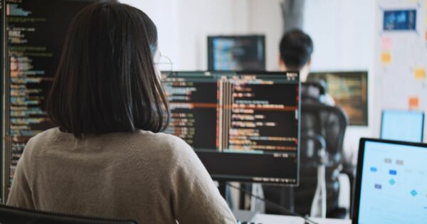 Uma mulher de cabelo escuro e óculos está sentada em frente a vários monitores de computador, trabalhando com código de programação. A tela principal exibe linhas de código coloridas em um ambiente de desenvolvimento. No fundo, outro programador, um homem de cabelo curto, também trabalha em seu computador, e há um quadro branco com anotações e diagramas. O ambiente parece ser um escritório moderno de tecnologia ou uma startup. (contrata, Prompting, dev)