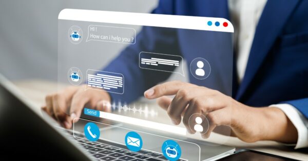 A imagem mostra uma pessoa vestindo um terno azul e utilizando um laptop. Sobre a tela do computador, há uma interface digital holográfica com elementos de um chatbot de atendimento ao cliente. A interface exibe ícones de mensagens, perfis de usuários e símbolos de comunicação, como telefone e e-mail, além de um balão de fala com a mensagem "HI! How can I help you?" (criação, atendimento)