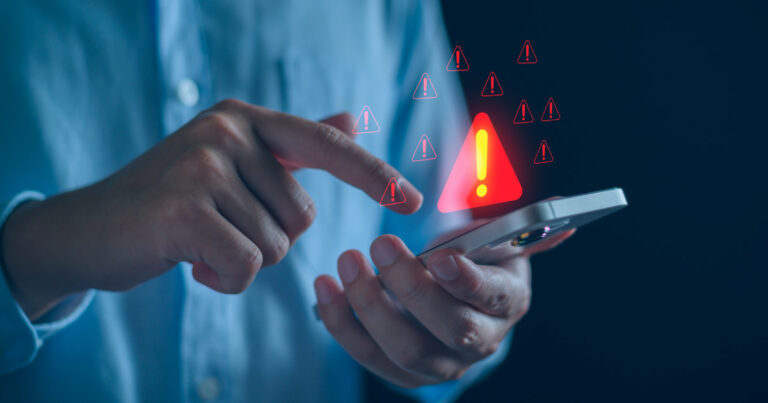 A imagem mostra uma pessoa segurando um smartphone enquanto toca na tela com o outro dedo. Diversos ícones vermelhos de alerta com pontos de exclamação aparecem sobrepostos à cena, simbolizando um aviso ou risco relacionado ao uso do dispositivo. O fundo escuro e desfocado destaca a interação e transmite uma sensação de preocupação ou atenção necessária, sugerindo um possível problema de segurança digital, golpe ou notificação de erro (avisamos, ataques, golpes, Brasil, Gartner)