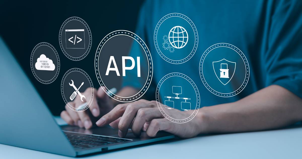 Pessoa usando um notebook enquanto diversos ícones digitais relacionados a tecnologia e segurança de APIs flutuam na imagem, incluindo símbolos de programação, nuvem de dados, ferramentas de configuração, globo representando internet, fluxogramas e um escudo com cadeado. A palavra "API" aparece em destaque no centro (ouribank)