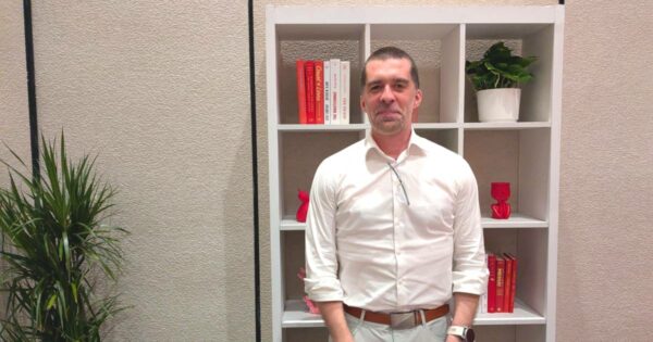 Homem em pé, usando camisa social branca e calça clara, com óculos pendurado na gola da camisa. Ao fundo, há uma estante branca com livros, objetos decorativos vermelhos e um vaso de planta. À esquerda da imagem, há uma planta decorativa. Trata-se de Mat Velloso, VP de Produto - Plataforma de Desenvolvimento em IA do Google DeepMind