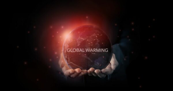 Imagem simbólica de duas mãos a segurar um globo terrestre iluminado por uma luz vermelha intensa, representando o aquecimento global. No centro do planeta, em destaque, aparece o texto 'GLOBAL WARMING'. O fundo é escuro, com pequenas partículas luminosas espalhadas, sugerindo calor ou partículas no espaço.