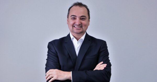 Homem de fato escuro e camisa branca sorridente, de braços cruzados, posando contra fundo cinzento. ingram