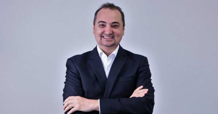 Homem de fato escuro e camisa branca sorridente, de braços cruzados, posando contra fundo cinzento. ingram