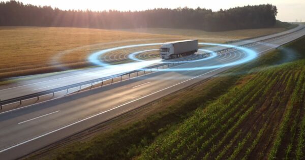 Um caminhão branco trafega em uma rodovia cercada por campos verdes e dourados ao pôr do sol. Há um efeito gráfico azul simulando ondas ao redor do veículo, sugerindo tecnologia de sensores ou conectividade digital na estrada (cimento, reduzir)