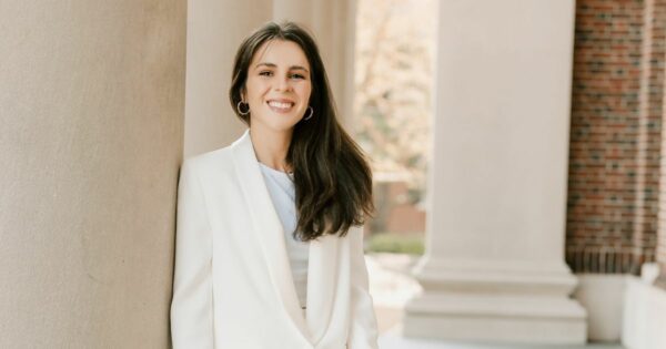 Mulher jovem a sorrir, vestida com blazer branco, encostada a uma coluna de pedra num espaço exterior com arquitetura clássica ao fundo.