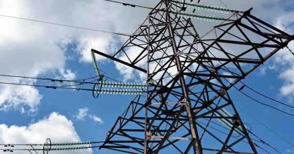 Fotografia de uma torre metálica de transmissão de energia elétrica vista de baixo contra um céu azul com algumas nuvens brancas. A estrutura de ferro é composta por uma malha de vigas diagonais e verticais, suportando cabos de alta tensão conectados através de isoladores de vidro verde-azulado em série (Inscrições, axia, equatorial)
