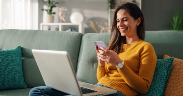 Mulher sorridente sentada em um sofá verde, usando um smartphone enquanto também está com um notebook no colo. Ela veste um suéter amarelo e parece estar em um ambiente doméstico moderno e aconchegante, sugerindo o uso da internet ou serviços digitais via smartphone em um momento de lazer ou trabalho remoto (brasil, colaboradores, híbrido)