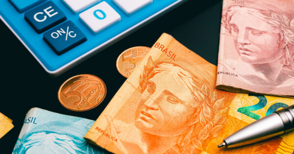 Imagem com cédulas e moedas de real, moeda oficial do Brasil, dispostas sobre uma superfície preta. Ao lado das cédulas e moedas, há uma calculadora azul e branca e uma caneta metálica prateada (reforma tributária, governo)