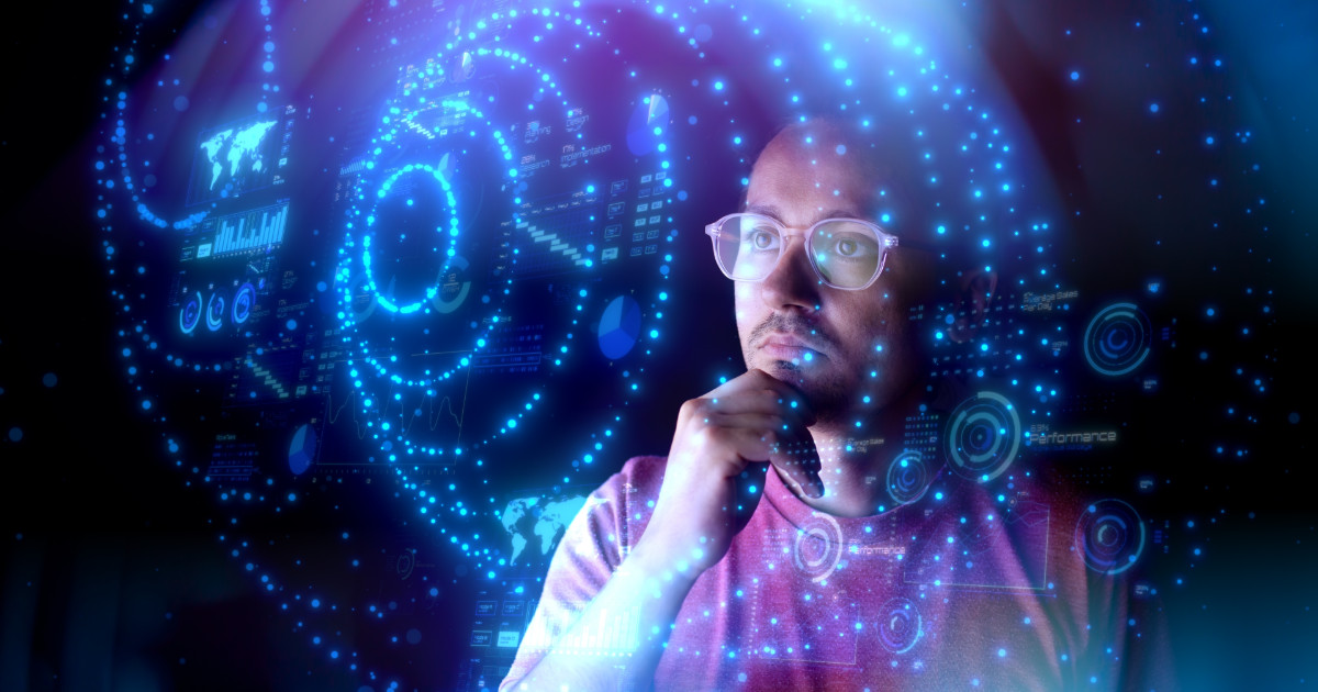 Homem usando óculos e camiseta vermelha observa atentamente uma interface digital futurista com gráficos, mapas e dados em azul, representando conceitos de inteligência artificial e análise de dados. A imagem transmite um ambiente tecnológico e inovador, relacionado a tendências em IA generativa (verity)