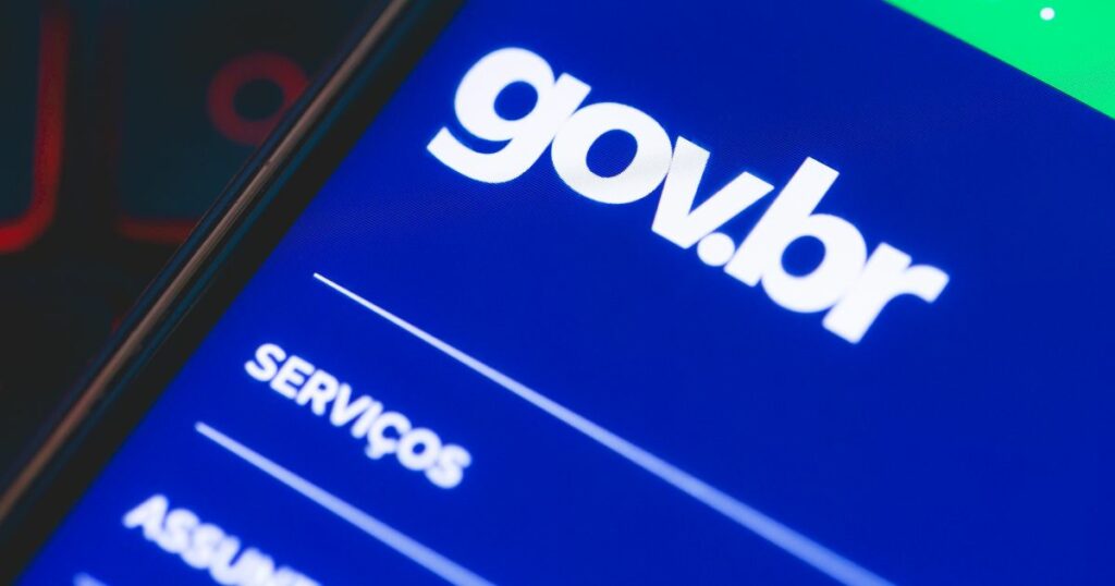 Imagem em close-up do ecrã de um smartphone a exibir a interface do portal digital do governo brasileiro, com o logótipo “gov.br” em letras brancas sobre um fundo azul. Abaixo, vê-se parcialmente a palavra “SERVIÇOS”, sugerindo a navegação por opções disponíveis no site. A imagem transmite a ideia de acesso digital a serviços, segurança, públicos, gargalos (suse)