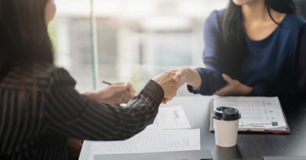 Duas mulheres apertam as mãos numa sala de reuniões, sugerindo o encerramento positivo de uma entrevista para uma vaga de emprego. Sobre a mesa estão documentos, incluindo um currículo, um contrato e um café para levar. A cena transmite profissionalismo e formalidade.