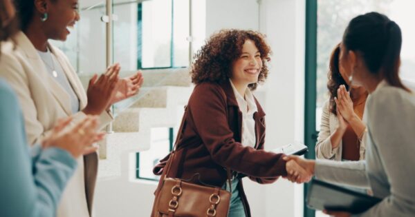 Mulher sorridente a apertar a mão a outra mulher num ambiente de escritório de Tecnologia, enquanto outras colegas aplaudem ao fundo, demonstrando boas-vindas ou celebração profissional.