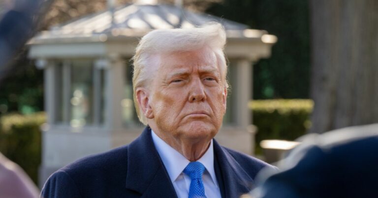 Um homem de expressão séria, com cabelo loiro penteado para trás e pele com tonalidade bronzeada, veste um sobretudo escuro, camisa branca e gravata azul. Ele está ao ar livre, em frente a uma estrutura clara com janelas, possivelmente parte de um edifício governamental ou um jardim formal. O fundo é composto por vegetação e arquitetura clássica, sugerindo um ambiente institucional (donald trump, eua, produtos)