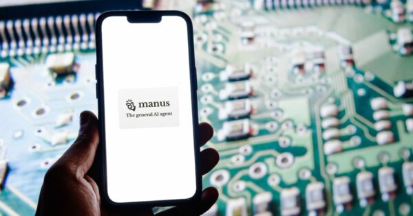 Mão segurando um smartphone com o ecrã a exibir o logótipo da "Manus – The general AI agent", sobre um fundo branco. Ao fundo, desfocado, vê-se um circuito eletrónico verde, repleto de componentes, simbolizando tecnologia e agente de inteligência artificial (agente)